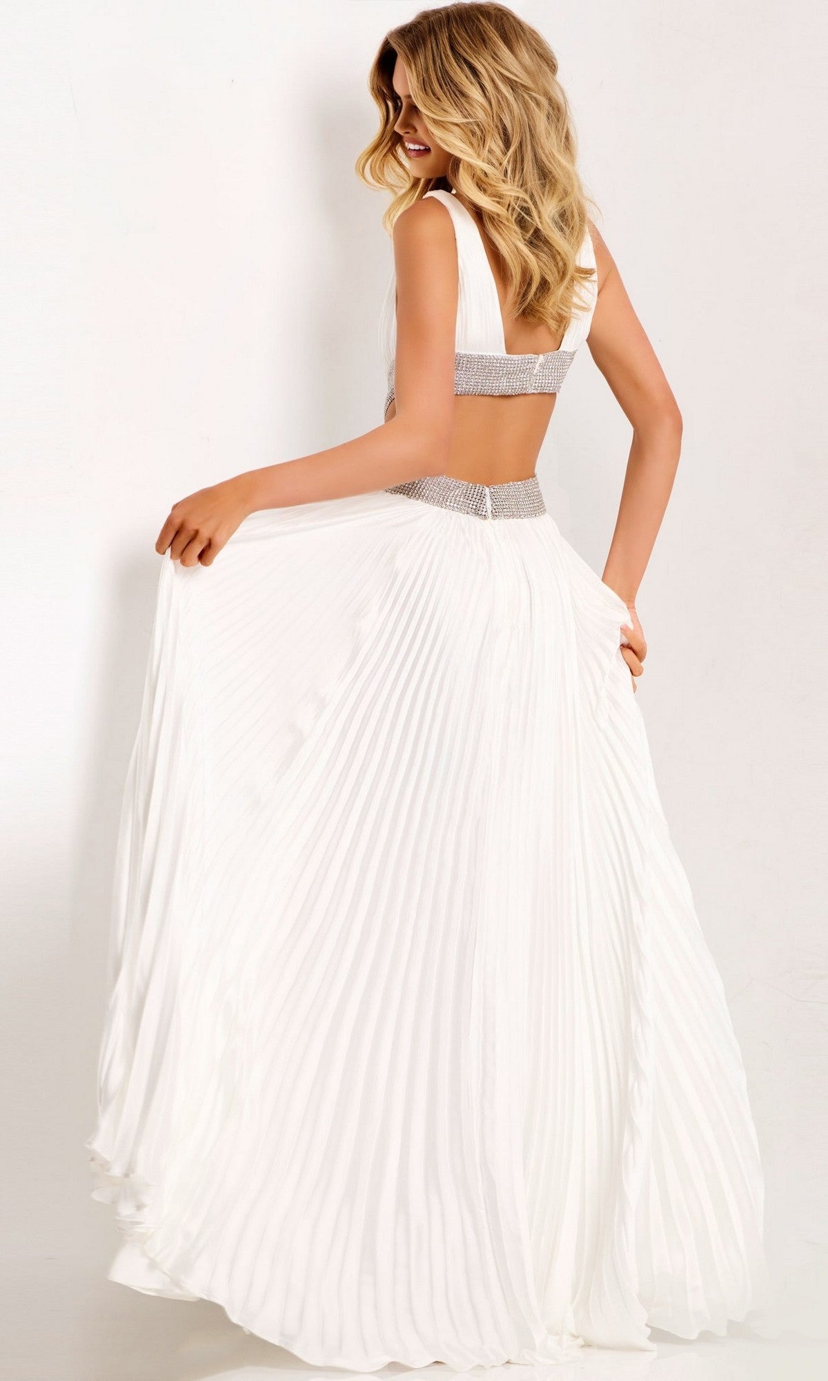 A woman models the Jovani 42811 long prom dress—sleeveless chiffon with an embellished waistband and silver bands at the waist and back—standing against a plain backdrop and glancing over her shoulder.
